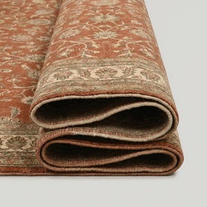 May include: Rolled-up area rug with a detailed floral pattern in shades of rust, beige, and green. The rug's edges are visible, showcasing the intricate design and texture. The rug is partially rolled, revealing its layered construction.