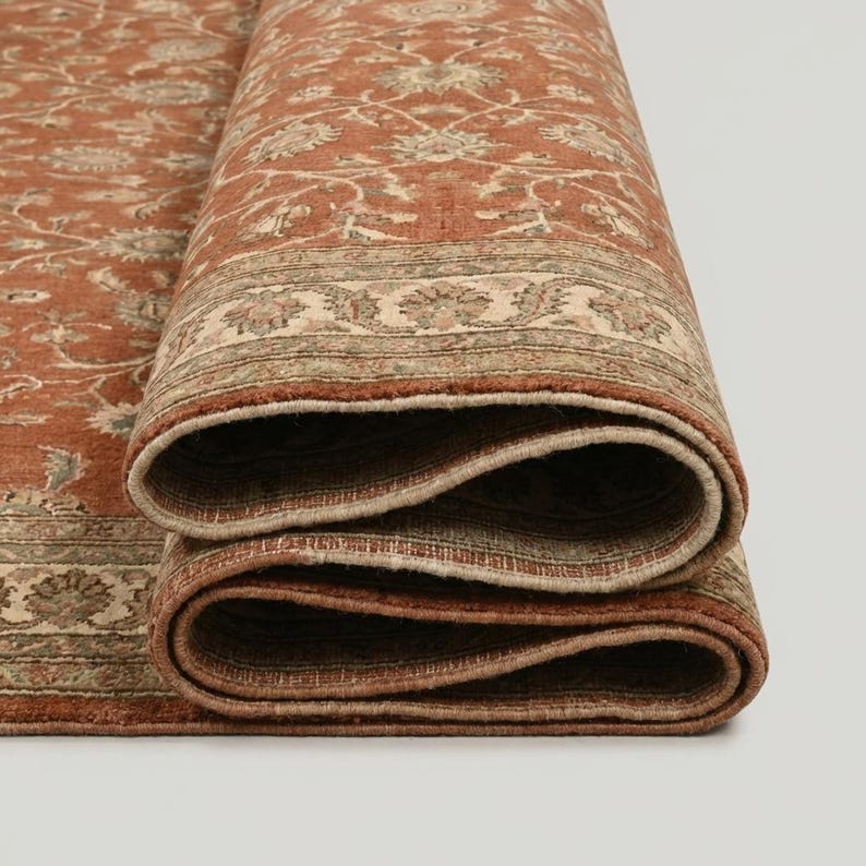 May include: Rolled-up area rug with a detailed floral pattern in shades of rust, beige, and green. The rug's edges are visible, showcasing the intricate design and texture. The rug is partially rolled, revealing its layered construction.