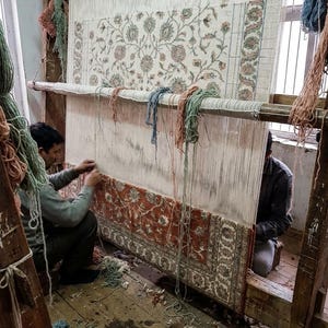 May include: Two people weaving a large rug on a wooden loom. The rug features a floral pattern in shades of red, cream, and green. The loom is set up in a room with wooden floors and natural light.