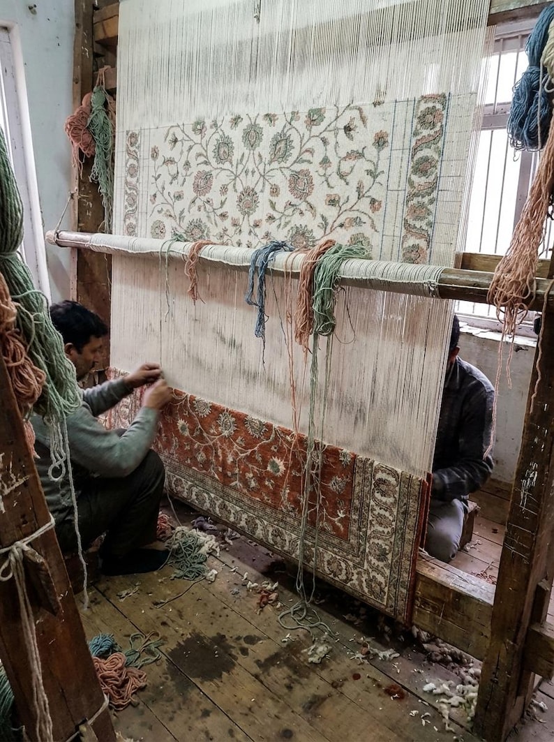 May include: Two people weaving a large rug on a wooden loom. The rug features a floral pattern in shades of red, cream, and green. The loom is set up in a room with wooden floors and natural light.