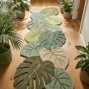 May include: A long runner rug featuring a series of overlapping monstera leaf shapes in various shades of green, sage, and cream. The rug has a textured, tufted surface with carved details outlining the veins of each individual leaf design.