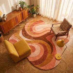 May include: A living room scene with a retro design. A large, abstract rug in peach, orange, and brown tones is the focal point. A mustard-yellow armchair and a patterned armchair are placed on the rug. A wooden cabinet and a yellow lamp are also visible.