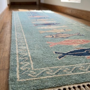 K&ouml;nnte beinhalten: Ein rechteckiger, hellblauer Teppich mit einem sich wiederholenden Fischmuster in Rosa, Blau und Beige. Der Teppich hat einen wei&szlig;en Rand mit einem dekorativen Design und Fransen an den Enden. Er liegt auf einem Holzboden.
