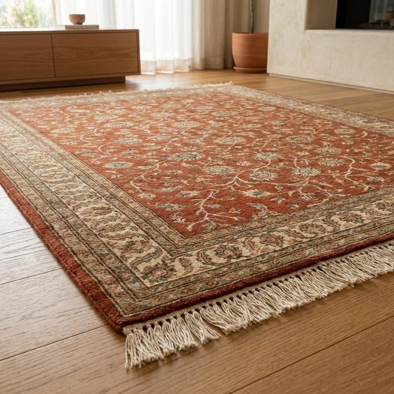 May include: A rectangular, rust-colored rug with an intricate floral pattern and a cream-colored border. The rug has a fringed edge and is placed on a hardwood floor. The design features a detailed, traditional aesthetic.