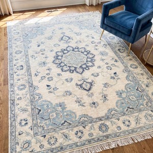 Op de afbeelding: Een rechthoekig vloerkleed met een crèmekleurige achtergrond en een gedetailleerd blauw bloemenpatroon. Het vloerkleed heeft een franje aan de randen en ligt op een houten vloer. Op de achtergrond is een blauwe fauteuil te zien.