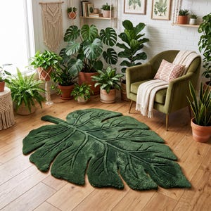 May include: A large, dark green area rug shaped like a monstera leaf with deep, textured veins and a soft, plush pile. The rug is positioned on a light-colored wooden floor, mimicking the organic, lobed silhouette of a tropical plant leaf.