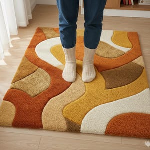 May include: A rectangular rug with a retro design featuring wavy patterns in shades of orange, brown, yellow, beige, and white. The rug has a textured, plush appearance. A person wearing blue jeans and white socks is standing on the rug.