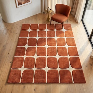 May include: A rectangular area rug featuring a repeating grid pattern of rounded, terracotta-colored rectangular shapes separated by thin, cream-colored lines. The rug has a soft, textured pile and covers a portion of the light-colored wooden floor.