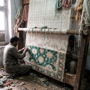 May include: Two people weaving a rug on a large wooden loom. The rug features a green and floral design. Various colored yarn bundles hang from the loom. The room has a rustic, aged appearance.