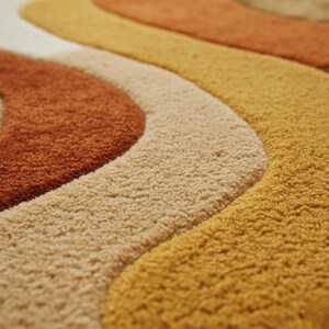 May include: Close-up of a retro-style rug with a wavy pattern. The rug features a textured surface and a color palette of orange, beige, and yellow. The design is reminiscent of the 1970s, with its bold, curved lines and warm tones.