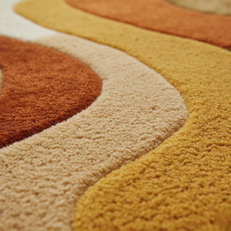 May include: Close-up of a retro-style rug with a wavy pattern. The rug features a textured surface and a color palette of orange, beige, and yellow. The design is reminiscent of the 1970s, with its bold, curved lines and warm tones.
