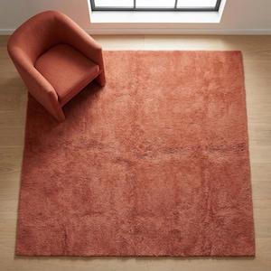 Puede incluir: Una alfombra de área de felpa de color terracota con un sillón a juego. La alfombra es rectangular y cubre una gran parte del suelo de madera. La silla tiene respaldo y reposabrazos curvos, cerca de una ventana.