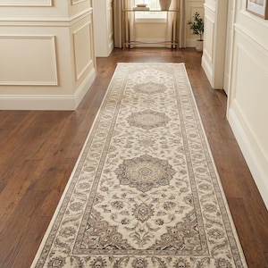 Peut inclure: Un long tapis de couloir rectangulaire présentant une base de couleur crème avec des motifs floraux et des médaillons symétriques complexes dans des tons taupe, gris et beige. Le tapis est fini avec des franges courtes et claires.