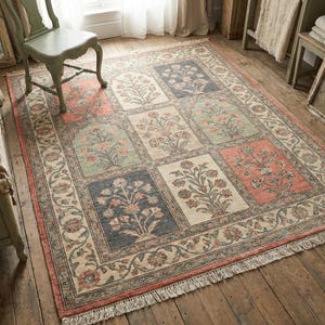 Op de afbeelding: Een rechthoekig vloerkleed met een bloemenpatroon. Het kleed heeft een herhalend ontwerp van bloemen vierkanten in beige, blauw en koraal, omlijst door een gedetailleerde rand. Het kleed heeft een gefranjerde rand en ligt op een houten vloer. Afmetingen in centimeters.