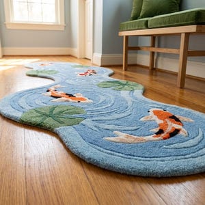 Puede incluir: Una alfombra azul con forma de agua con un dise&ntilde;o de estanque de carpas koi. La alfombra presenta peces koi en naranja, negro y blanco, junto con nen&uacute;fares verdes. La alfombra est&aacute; colocada sobre un suelo de madera.