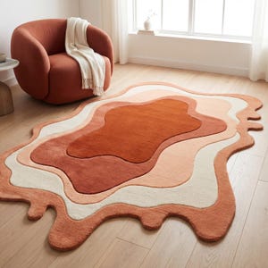 Op de afbeelding: Een abstract tapijt met een gelaagd ontwerp in tinten oranje, perzik en crème. Het tapijt heeft een onregelmatige, vloeiende vorm en voegt een moderne touch toe. Het tapijt ligt op een houten vloer. Afmetingen in centimeters.