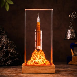May include: A rectangular resin block containing a miniature model of a NASA rocket with orange and white segments. The base of the rocket is surrounded by an illuminated, textured orange resin plume, and the entire block sits on a wooden base.