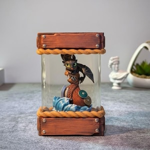 Könnte beinhalten: Ein dekoratives Glasgefäß mit Holzrahmen und Seilbesatz. Im Inneren sitzt eine Figur eines Drachencharakters auf einem stilisierten Boot mit blauen Wellen. Der Drache ist schwarz mit grünen Augen und Flügeln.