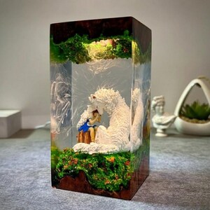 May include: A rectangular resin lamp featuring a scene with a white wolf and a girl in a blue dress. The lamp has a wooden base and is filled with green foliage and a light source, creating a warm glow. The lamp is a decorative item.