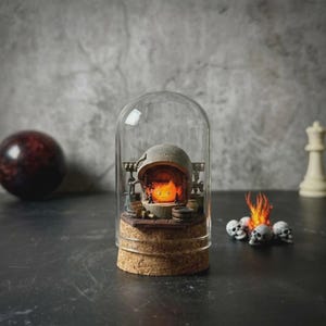 May include: A miniature diorama enclosed in a glass dome with a cork base, featuring a small orange flame character inside a stone hearth structure. Beside the dome, a small cluster of white skulls surrounds a flickering orange flame figurine.