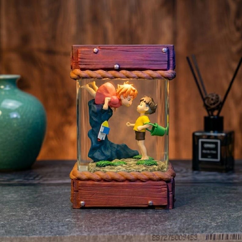 K&ouml;nnte beinhalten: Dekorative Glasbox mit einer Szene von zwei Zeichentrickfiguren. Eine ist kopf&uuml;ber, die andere h&auml;lt einen gr&uuml;nen Eimer. Die Box hat einen Holzrahmen mit Seildetails. Sie steht auf einer dunklen Oberfl&auml;che, mit einer Vase und einem Diffusor im Hintergrund.