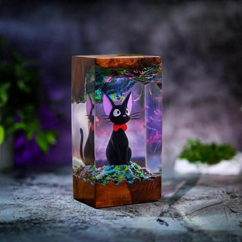 May include: A black cat figurine with a red bow tie sits inside a clear resin block. The resin is encased in a wooden frame, with colorful details visible within the resin. The cat has large eyes and pointed ears.