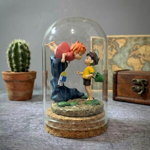 K&ouml;nnte beinhalten: Eine Glaskuppel auf einem Korksockel beherbergt eine Miniaturszene mit zwei animierten Figuren. Eine Figur mit roten Haaren und rotem Kleid lehnt sich &uuml;ber eine blaue Felsformation. Die andere Figur, in einem gelben Hemd und braunen Shorts, steht darunter. Links ein kleiner Kaktus in einem Terrakottatopf.