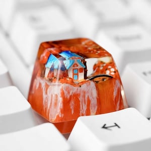 Può includere: Un copritasto artigianale personalizzato con una scena di casa in miniatura. Il copritasto è arancione con dettagli bianchi, e la casa ha un tetto blu e pareti bianche. Il copritasto è posto su una tastiera bianca.