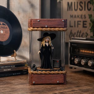 Könnte beinhalten: Eine dekorative Musik-Themen-Anzeige mit einer Miniaturfigur einer Sängerin in einem schwarzen Kleid und Hut, die ein Mikrofon hält. Die Figur befindet sich in einem Glaskasten mit Holzrahmen und Seilakzenten. Ein kleiner Verstärker ist ebenfalls vorhanden.