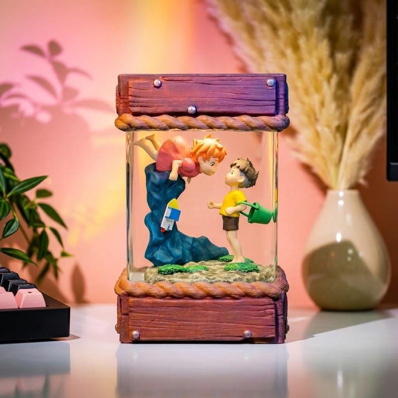K&ouml;nnte beinhalten: Dekorative Glasfigur mit zwei animierten Figuren. Eine Figur schwebt &uuml;ber einer blauen Felsformation, w&auml;hrend die andere auf einer sandigen Basis steht und eine gr&uuml;ne Gie&szlig;kanne h&auml;lt. Die Figur ist in einem Holzrahmen eingeschlossen.