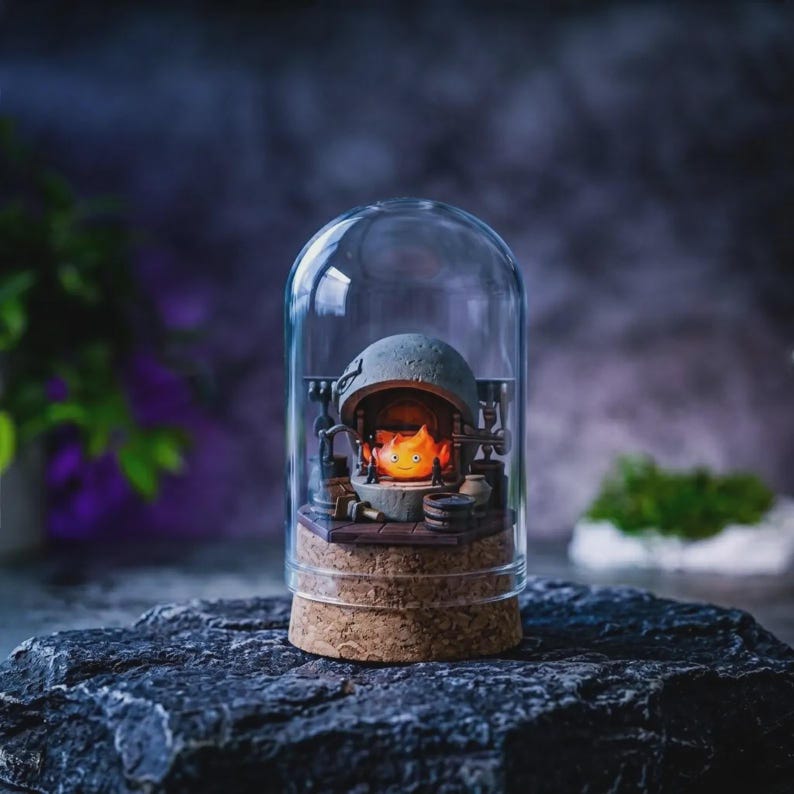 May include: A clear glass dome cloche with a cork base containing a miniature diorama of a stone hearth. Inside the hearth sits a small, glowing orange and yellow flame character with a smiling face, surrounded by tiny dark gray tools and accessories.
