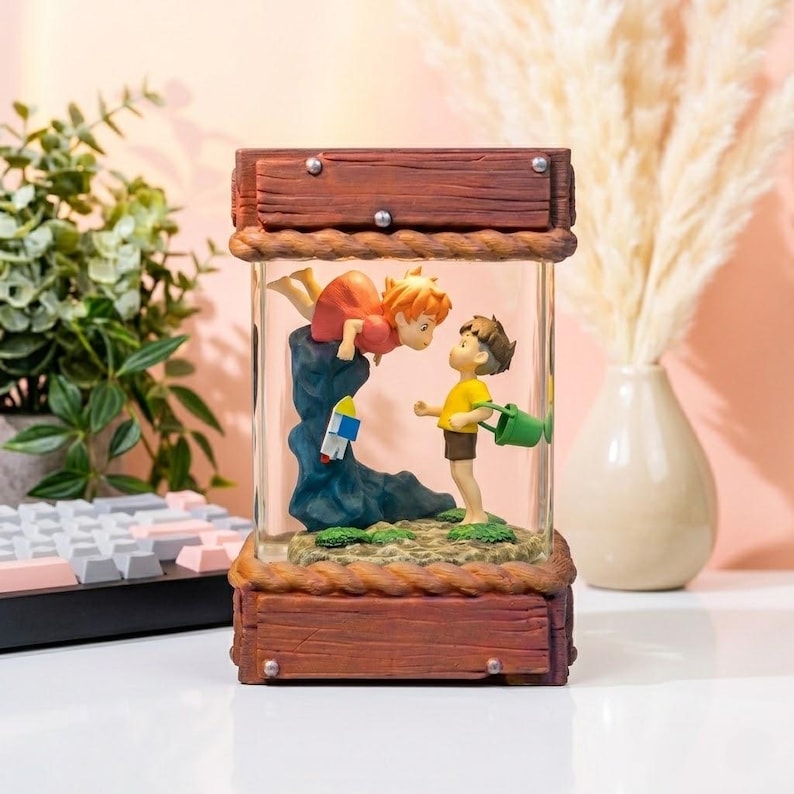 K&ouml;nnte beinhalten: Eine dekorative Glasbox mit einer Szene mit zwei Zeichentrickfiguren. Eine Figur schwebt in der Luft, die andere steht auf einem kleinen Grasb&uuml;schel. Die Box hat einen Holzrahmen und eine seilartige Umrandung.