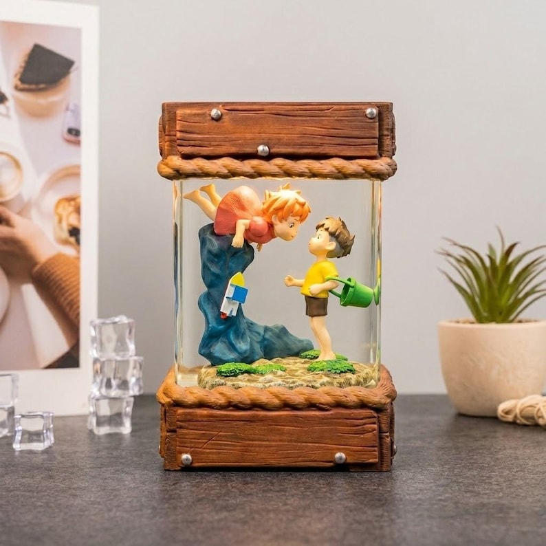 K&ouml;nnte beinhalten: Eine dekorative Glasbox mit Holzrahmen und Seilrand. Im Inneren sind zwei Zeichentrickfiguren dargestellt: ein M&auml;dchen in einem roten Kleid und ein Junge in einem gelben Hemd. Das M&auml;dchen befindet sich auf einem blauen Felsen, und der Junge h&auml;lt eine gr&uuml;ne Gie&szlig;kanne.