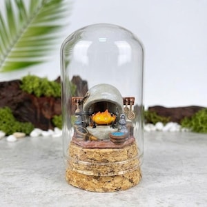 May include: A miniature diorama inside a clear glass dome with a cork base. The scene features a small orange flame character sitting inside a grey stone fireplace, surrounded by tiny kitchen utensils, a blue bowl, and a grey jug on a stone hearth.