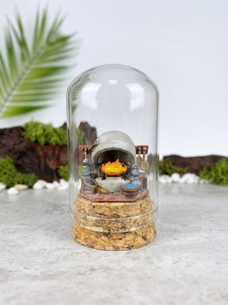 May include: A miniature diorama inside a clear glass dome with a cork base. The scene features a small orange flame character sitting inside a grey stone fireplace, surrounded by tiny kitchen utensils, a blue bowl, and a grey jug on a stone hearth.
