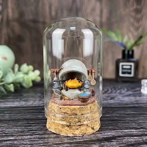 May include: A miniature diorama enclosed in a clear glass dome with a cork base. Inside, a small orange flame character sits within a grey stone fireplace. The scene includes tiny kitchen utensils, a blue bowl, and a grey kettle arranged around the hearth.