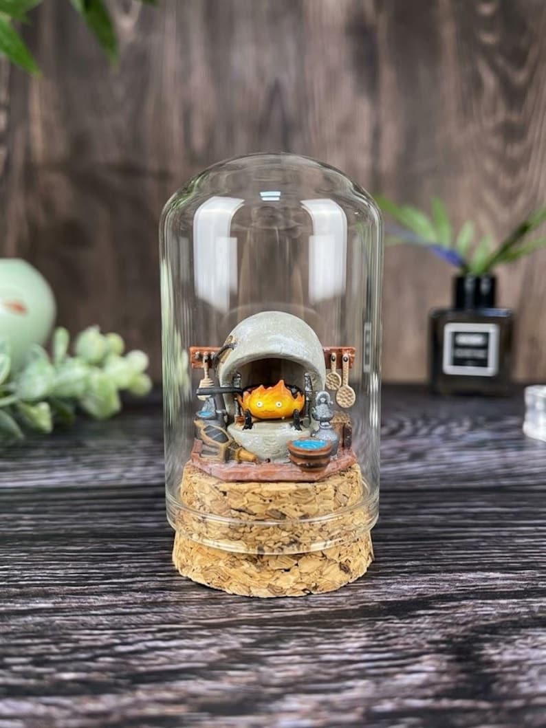 May include: A miniature diorama enclosed in a clear glass dome with a cork base. Inside, a small orange flame character sits within a grey stone fireplace. The scene includes tiny kitchen utensils, a blue bowl, and a grey kettle arranged around the hearth.