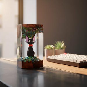May include: A black cat figurine with a red bow is encased in a clear resin block. The base is a dark wood, and the top has a colorful, textured design. The cat is sitting on a green and blue base. A unique decorative item.