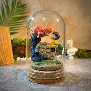 K&ouml;nnte beinhalten: Ein Glaskuppel-Terrarium mit einer Miniatur-Szene mit zwei Figuren im Anime-Stil. Eine Figur tr&auml;gt ein rotes Kleid, die andere ein gelbes Hemd und h&auml;lt eine gr&uuml;ne Gie&szlig;kanne. Die Basis ist aus Kork.