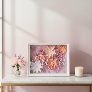 May include: A framed floral art piece with the name "June" in cursive script. The artwork features paper flowers in shades of pink, peach, and purple, set against a pink background. A small vase of pink flowers and a lit candle are nearby.