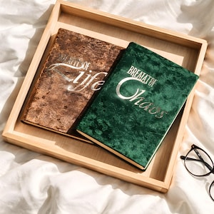 Könnte beinhalten: Zwei samtbezogene Journale, eines braun und eines grün, liegen in einem Holztablett. Das braune Journal trägt den silbernen Schriftzug "Breath of Life", während das grüne Journal "Breath of Chaos" zeigt. Eine Brille ist ebenfalls zu sehen.