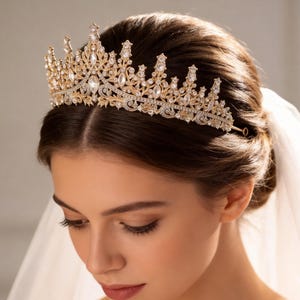 May include: A gold-toned bridal crown with clear crystals. The tiara features an ornate design with multiple peaks and a delicate, curved base. The crown is worn by a person with brown hair and a white veil.