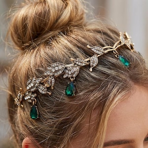 May include: A gold-toned hair crown with a leaf and crystal design. The crown features clear crystals and teardrop-shaped emerald green gems. The hair accessory is worn on a person with blonde hair.