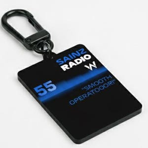 Könnte beinhalten: Schwarzer rechteckiger Schlüsselanhänger mit silbernem Verschluss. Der Schlüsselanhänger hat blauen Text mit der Aufschrift "SAINZ RADIO W", "55" und "SMOOTH OPERATOOOR!". Der Schlüsselanhänger befindet sich auf weißem Hintergrund.