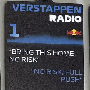 May include: Black rectangular keychain with the text "VERSTAPPEN RADIO" in white and blue. Features the number 1 and the phrases "BRING THIS HOME, NO RISK" and "NO RISK, FULL PUSH". Includes a black metal clasp.