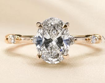 May include: A rose gold ring featuring a large oval-cut center stone held by four prongs. The band is accented with two smaller round-cut stones on each side, positioned near the center setting, and one additional round stone on each side of the band.