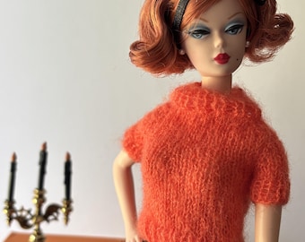 May include: A fashion doll with shoulder-length auburn hair styled in a flip, wearing a black headband with a bow, small pearl earrings, a bright orange fuzzy short-sleeved sweater, and a black tulle skirt, with a gold candelabra nearby.