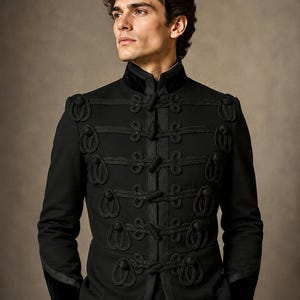 Könnte beinhalten: Eine schwarze Jacke mit hohem Stehkragen und einem Vorderverschluss, der mit fünf Reihen dekorativer schwarzer Kordelgeflechte und Knebelknöpfen verziert ist. Das Kleidungsstück hat strukturierte Schultern und lange Ärmel mit Samtbündchen.