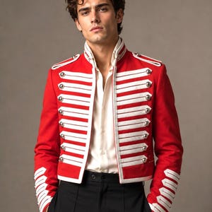 May include: A bright red military-style jacket featuring a high collar, white braided cord detailing across the chest, and silver-toned buttons. The jacket has structured shoulders with epaulets and white trim on the cuffs, worn over a white shirt.