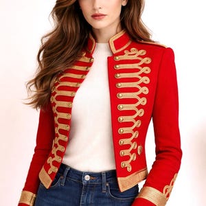 Puede incluir: Una chaqueta roja de estilo militar con detalles de cordón trenzado dorado, cuello alto y botones dorados en la parte delantera. La chaqueta incluye ribetes dorados en los puños y el dobladillo, sobre un top blanco acanalado y vaqueros azules.